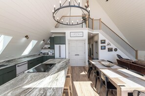 Eine geräumige Küche mit einer großen Kochinsel, ein Essbereich mit Holztischen, eine Treppe und ein Blick zum Himmel durch Dachfenster.
