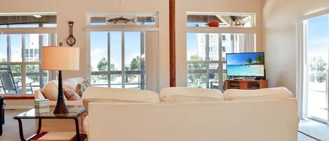 A living room with a sofa, a coffee table, a lamp, a TV, and large windows with a view of palm trees and buildings.