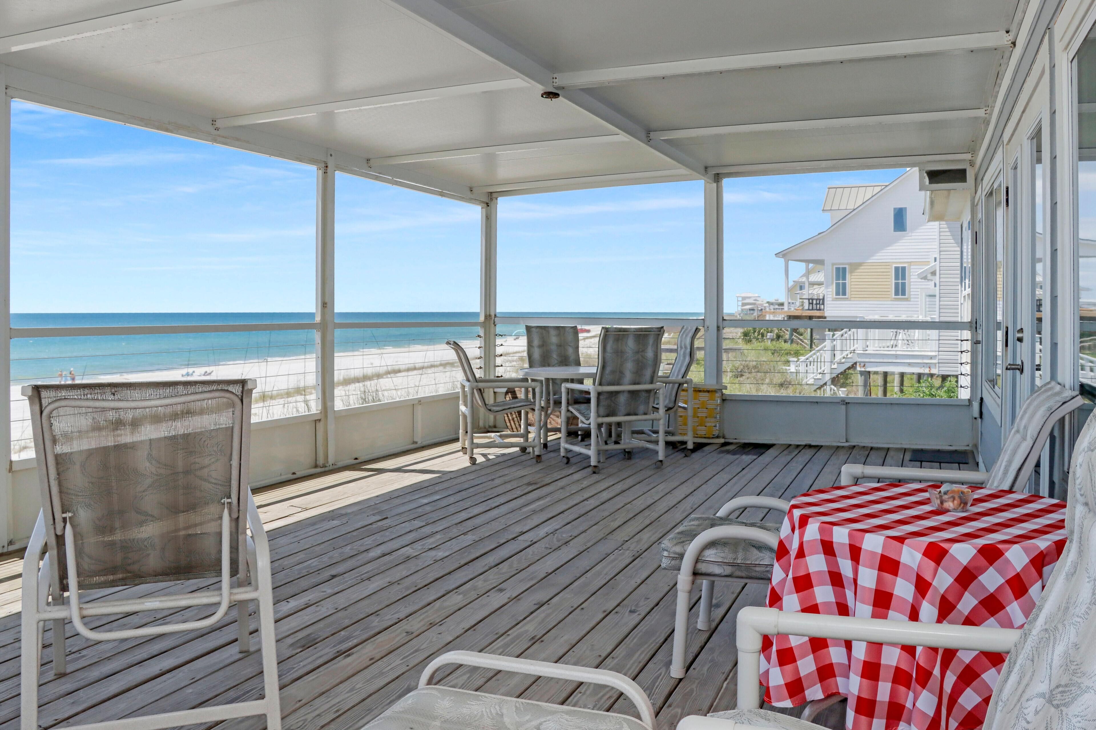 A covered deck with a view of the beach, a table with a checkered tablecloth, and a chair.