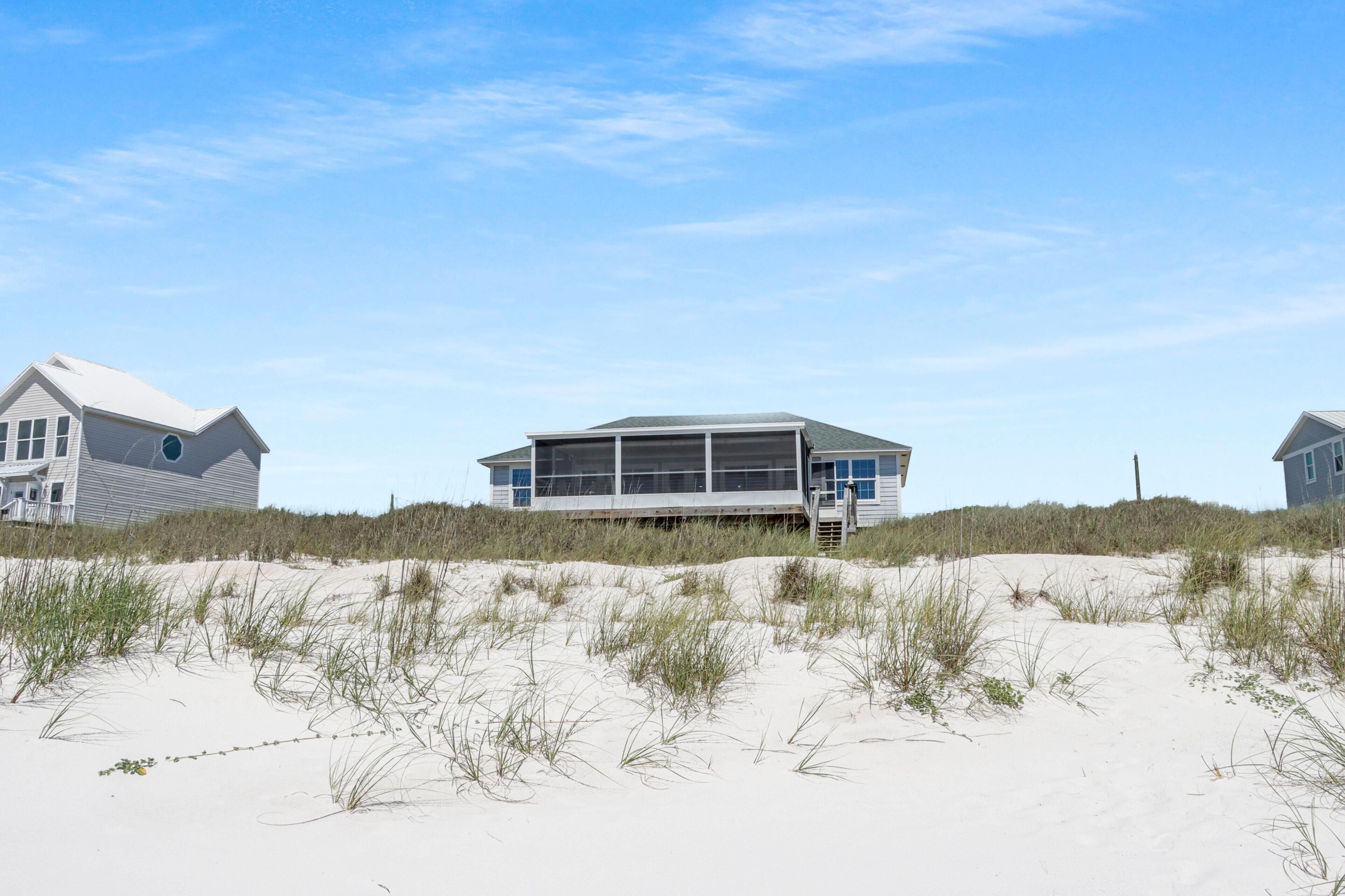 A beachfront house with a large windowed section, a covered porch, and a sandy beach in the foreground.
