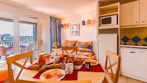 Una habitación con una mesa de comedor dispuesta con comida, un sofá con cojines de colores, una zona de cocina con electrodomésticos y vistas a través de una ventana.
