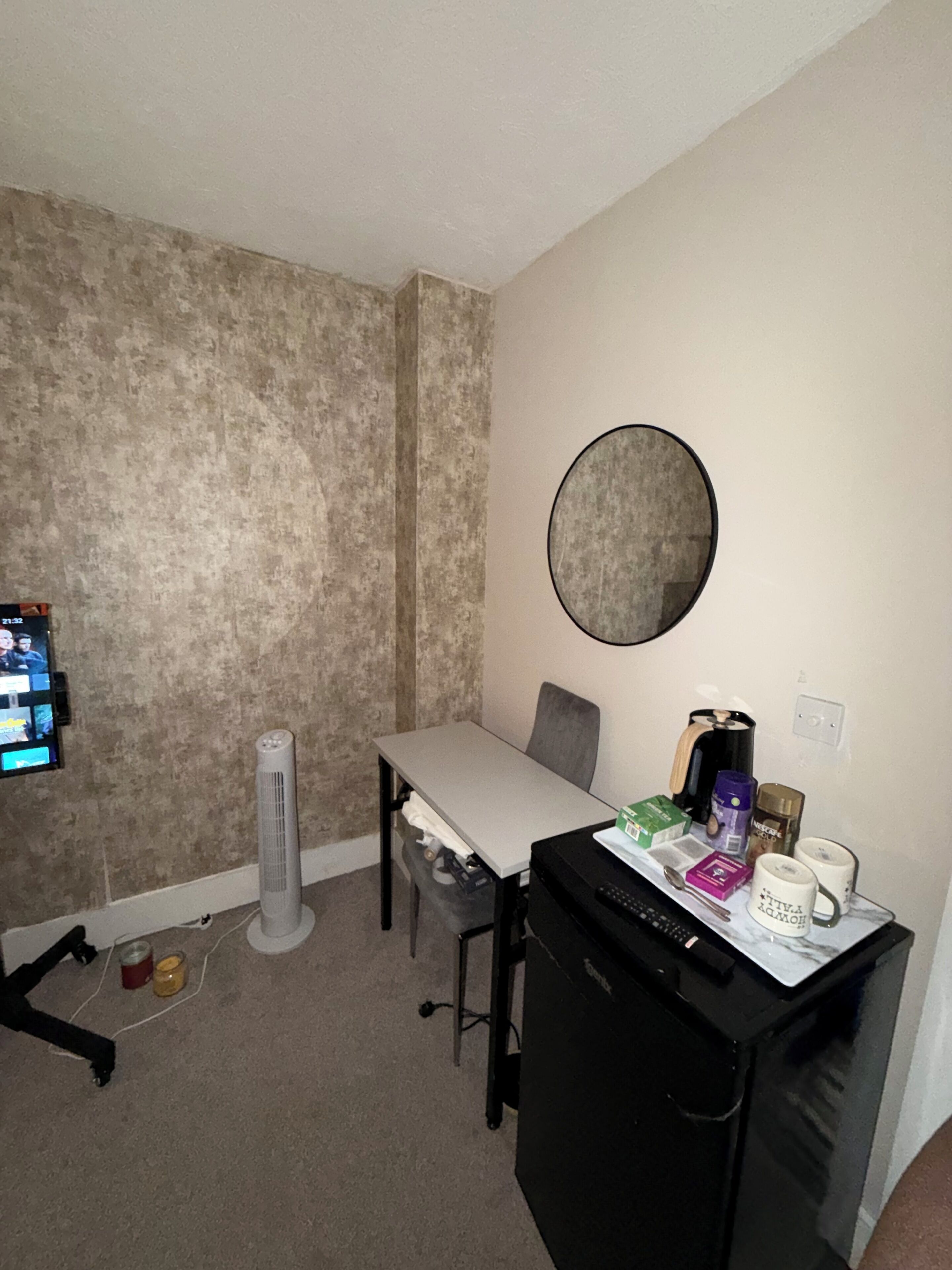 A small room with a white table, a chair, a black appliance, a round mirror, and a TV mounted on the wall.