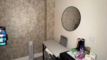 A small room with a white table, a chair, a black appliance, a round mirror, and a TV mounted on the wall.