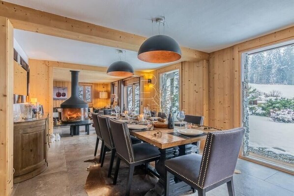 Ein Holzinterieur mit Esstisch, Stühlen, Pendelleuchten, einem Kamin und einem großen Fenster.