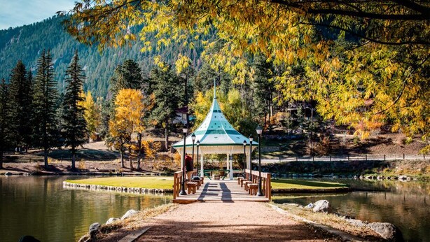 A gazebo with a green roof, surrounded by a lake, trees, and a mountain in the background.