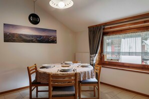 Un comedor con una mesa dispuesta para cuatro, sillas, un reloj, una ventana con cortinas y un cuadro de paisaje en la pared.