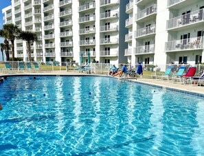 Ein Pool mit klarem, blauem Wasser, umgeben von einem weißen Gebäude mit Balkonen und einem eingezäunten Bereich mit Liegestühlen.