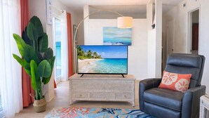 Un salon avec un téléviseur, un fauteuil en cuir noir, une grande plante et un tapis coloré.