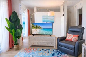 Ein Wohnzimmer mit Fernseher, einem schwarzen Ledersessel, einer großen Pflanze und einem farbenfrohen Teppich.