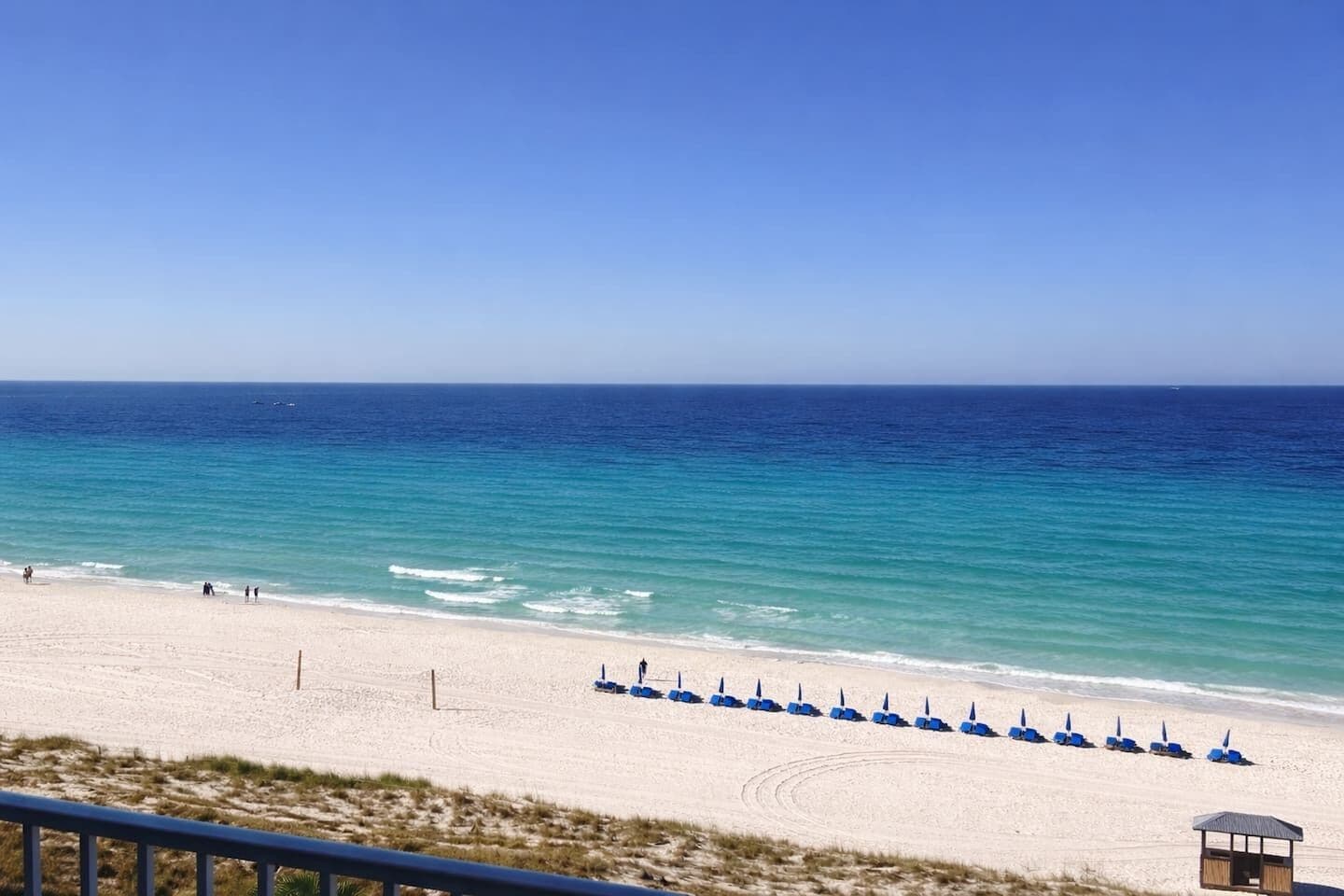 A beach with a row of blue umbrellas, a sandy shore, and a clear blue sky.