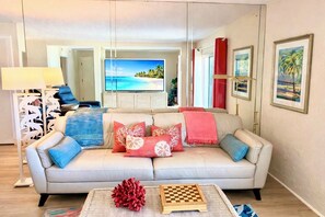 Ein Wohnzimmer mit einer Couch, einem Couchtisch mit Schachbrett und einem Fernseher, der eine Strandansicht zeigt.