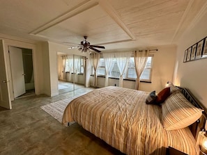 A bedroom with a large bed, a ceiling fan, a window with curtains, and a door leading to another room.
