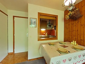Ein Essbereich mit einem Tisch für vier Personen, einer Holzwand und einer durch ein Fenster sichtbaren Küche.