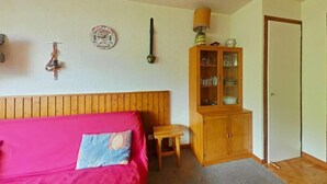 Quarto com cabeceira de madeira, cama com cobertura rosa, mesa de madeira pequena, aparador com copos e uma porta.