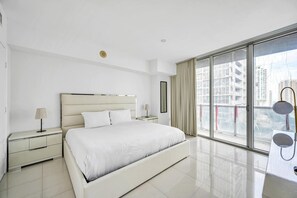 Ein modernes Hotelzimmer mit einem großen Bett, einem Nachttisch, einer Lampe, einem Fenster mit Blick auf Gebäude und einem Balkon mit Geländer.