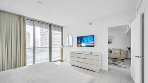 Moderni hotellihuone, jossa on suuri sänky, seinälle asennettu televisio, lasiliukuovi parvekkeelle ja näkymä kaupungin ylle.