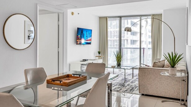 Moderni hotellihuone, jossa on lasinen ruokapöytä, sohva, televisio ja suuri ikkuna, jossa on verhot.
