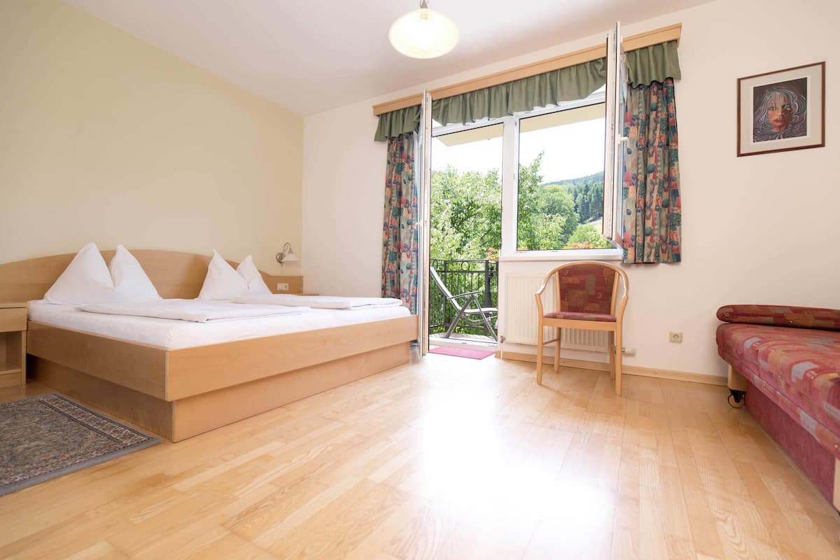 A bedroom with a bed, a chair, a sofa, a window with curtains, and a view of greenery outside.