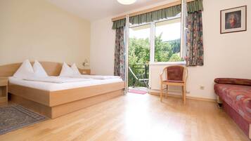 A bedroom with a bed, a chair, a sofa, a window with curtains, and a view of greenery outside.