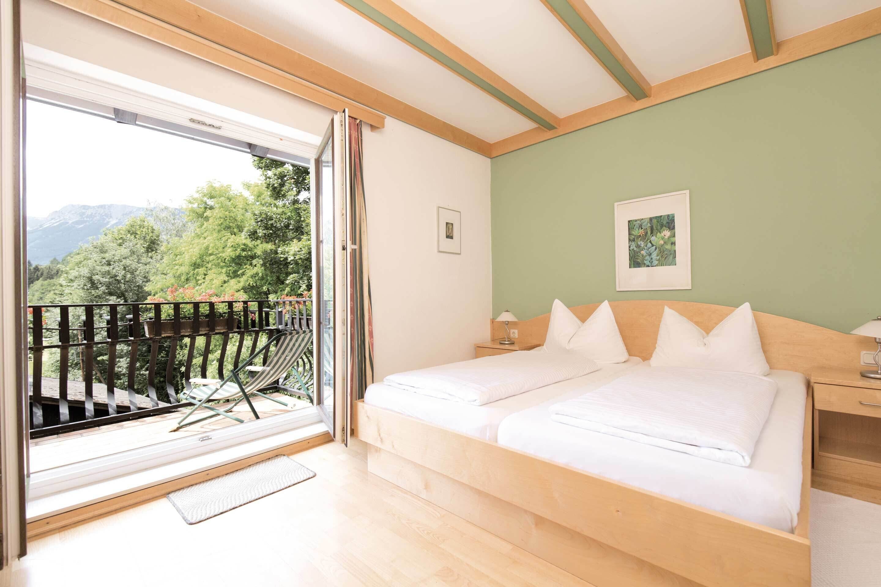 A bedroom with a bed, a nightstand, a window with a view of greenery, and a balcony with a chair.