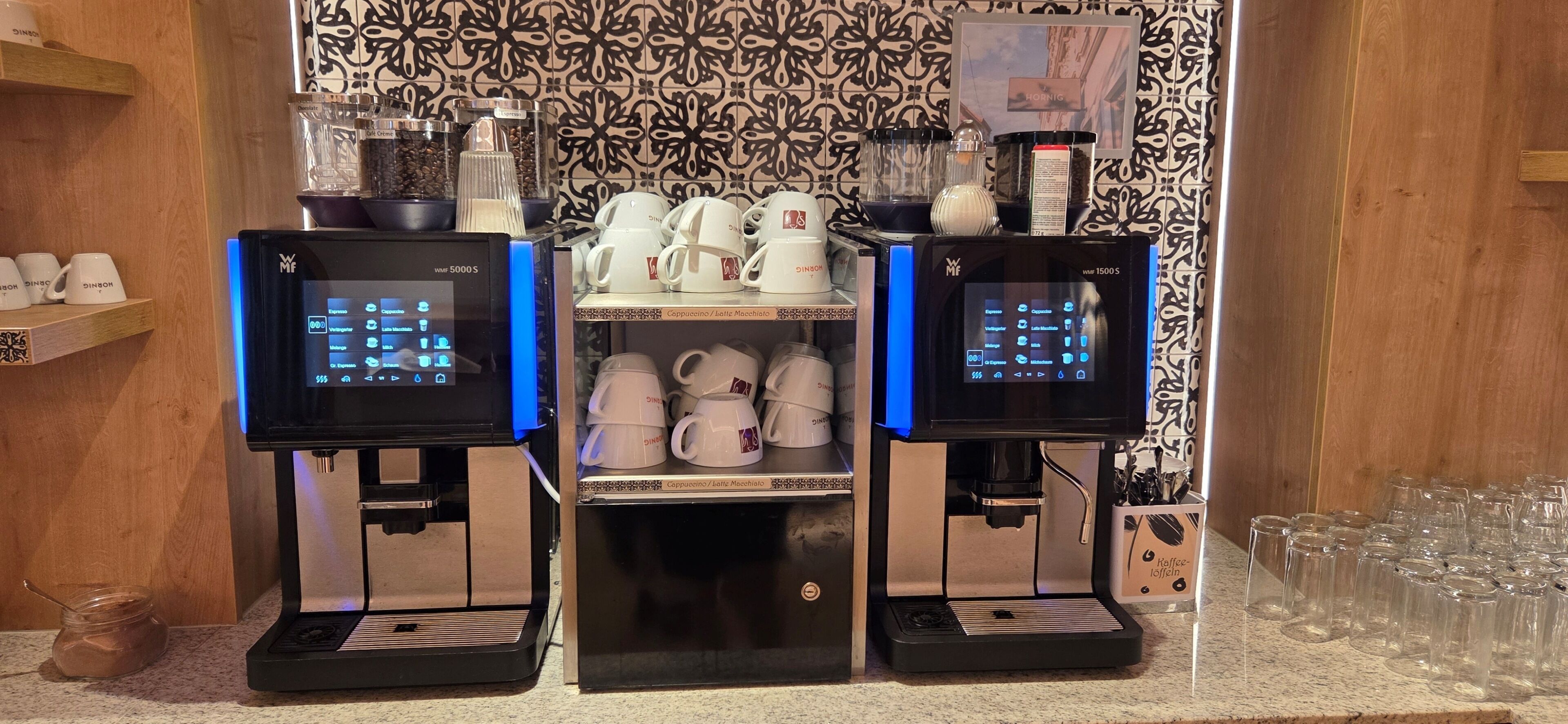 Dues màquines de cafè amb pantalles digitals, una estant amb tasses blanques i una paret amb un disseny decoratiu.