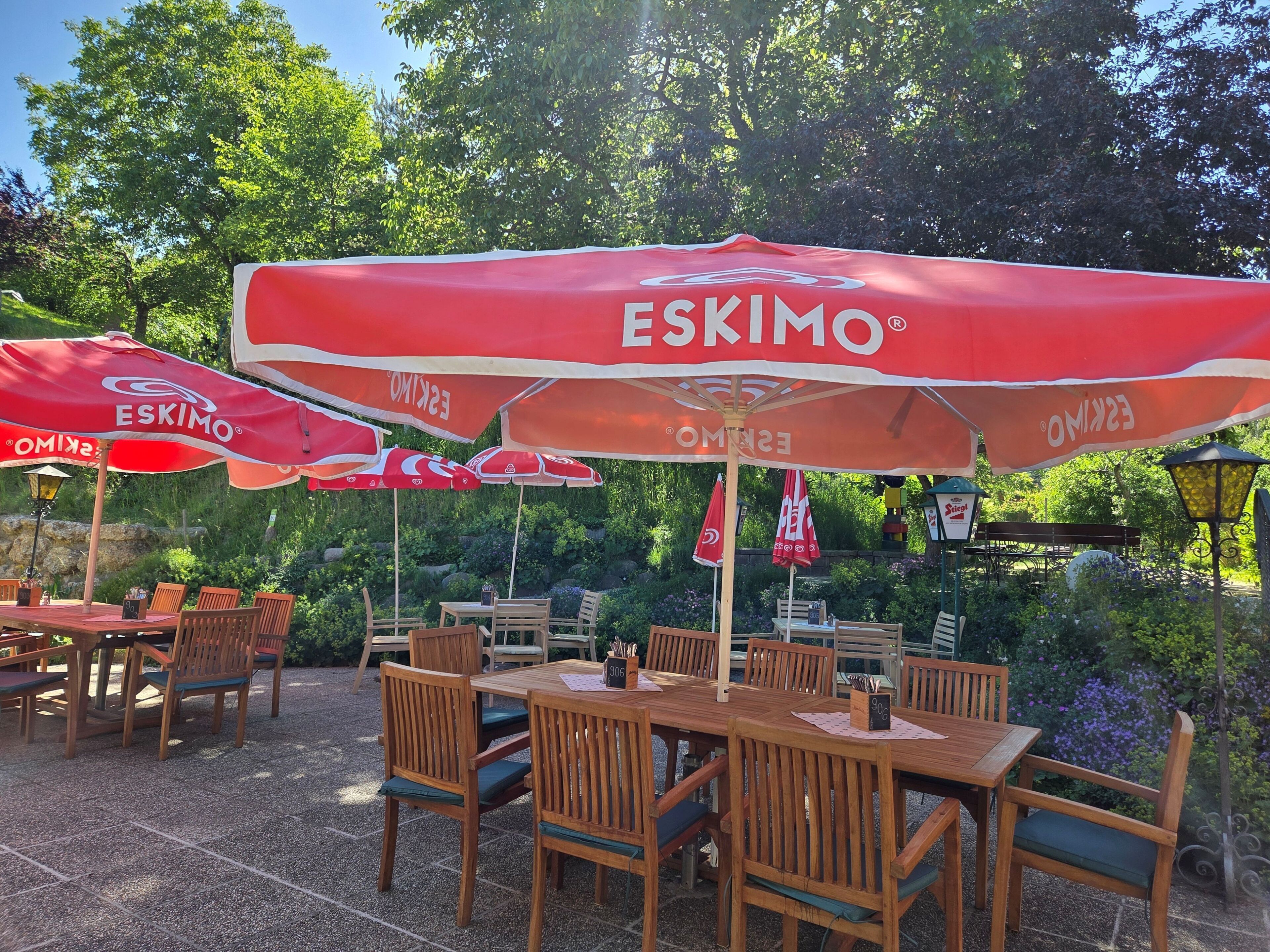 Mesas y sillas de madera al aire libre con sombrillas rojas, mostrando el nombre 'ESKIMO' en un jardín.