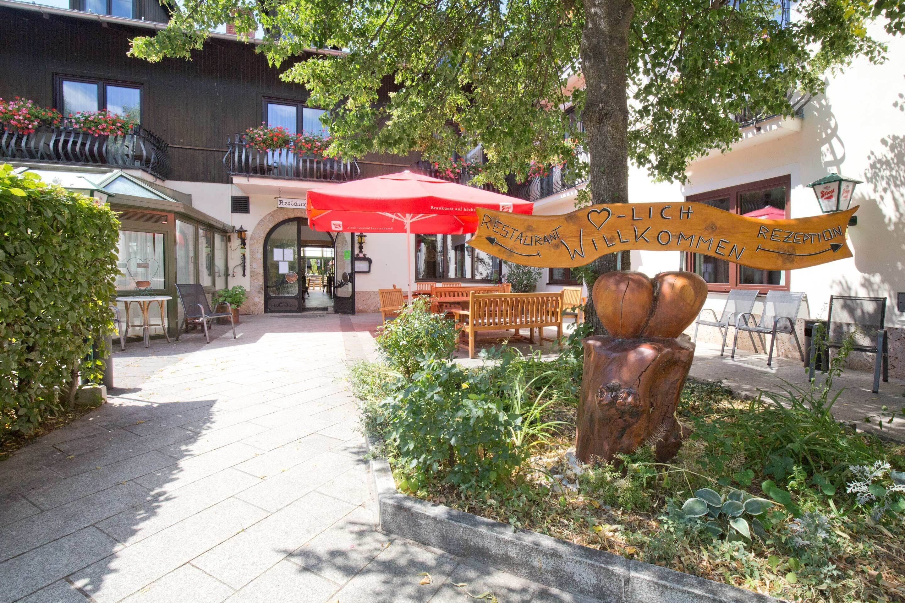 A courtyard with outdoor seating, a wooden sign with 'RESTAURANT' and 'WILLKOMMEN' text, and a red umbrella.