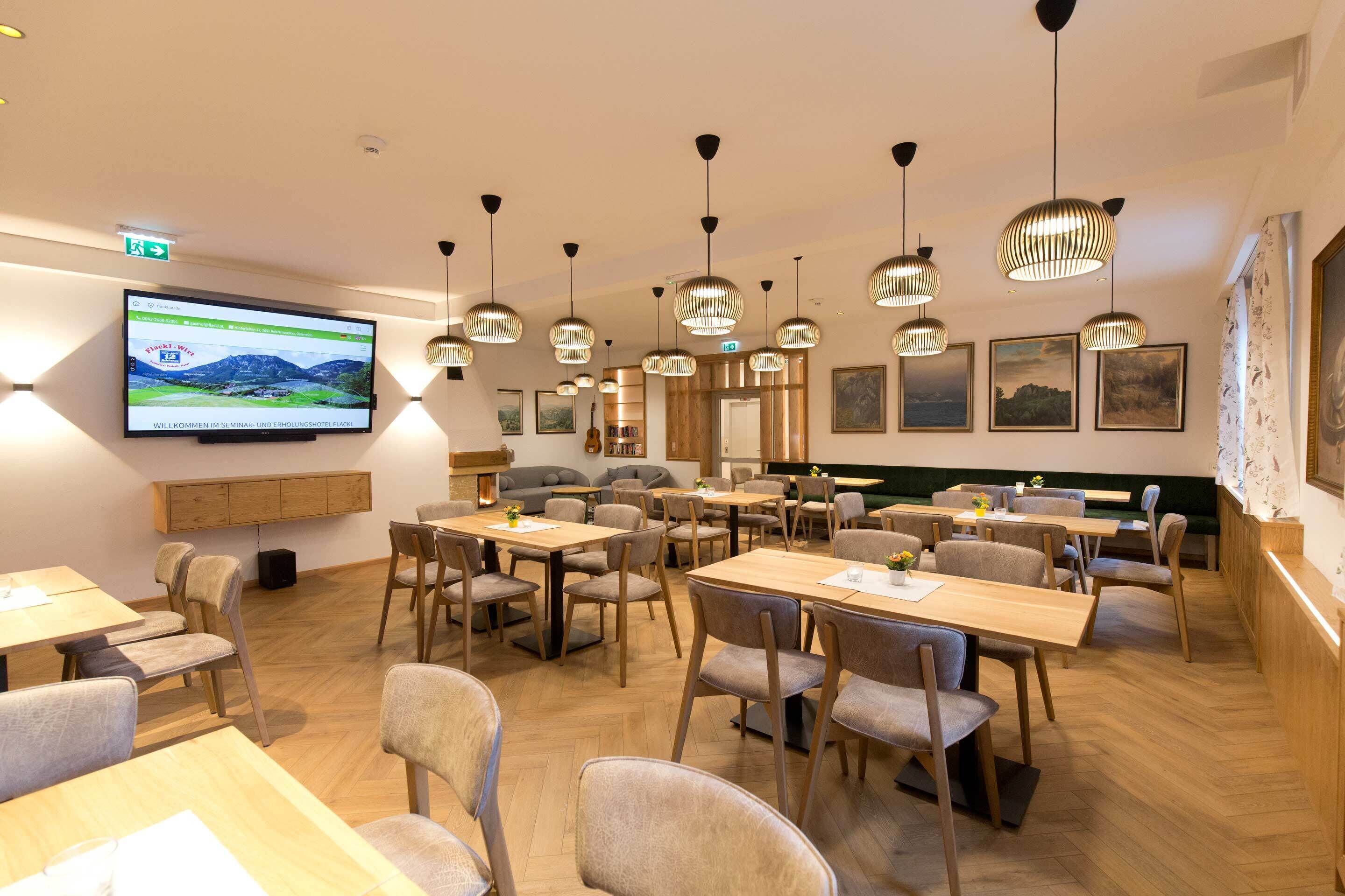 A restaurant with wooden tables and chairs, hanging pendant lights, a television screen, and framed artwork on the walls.