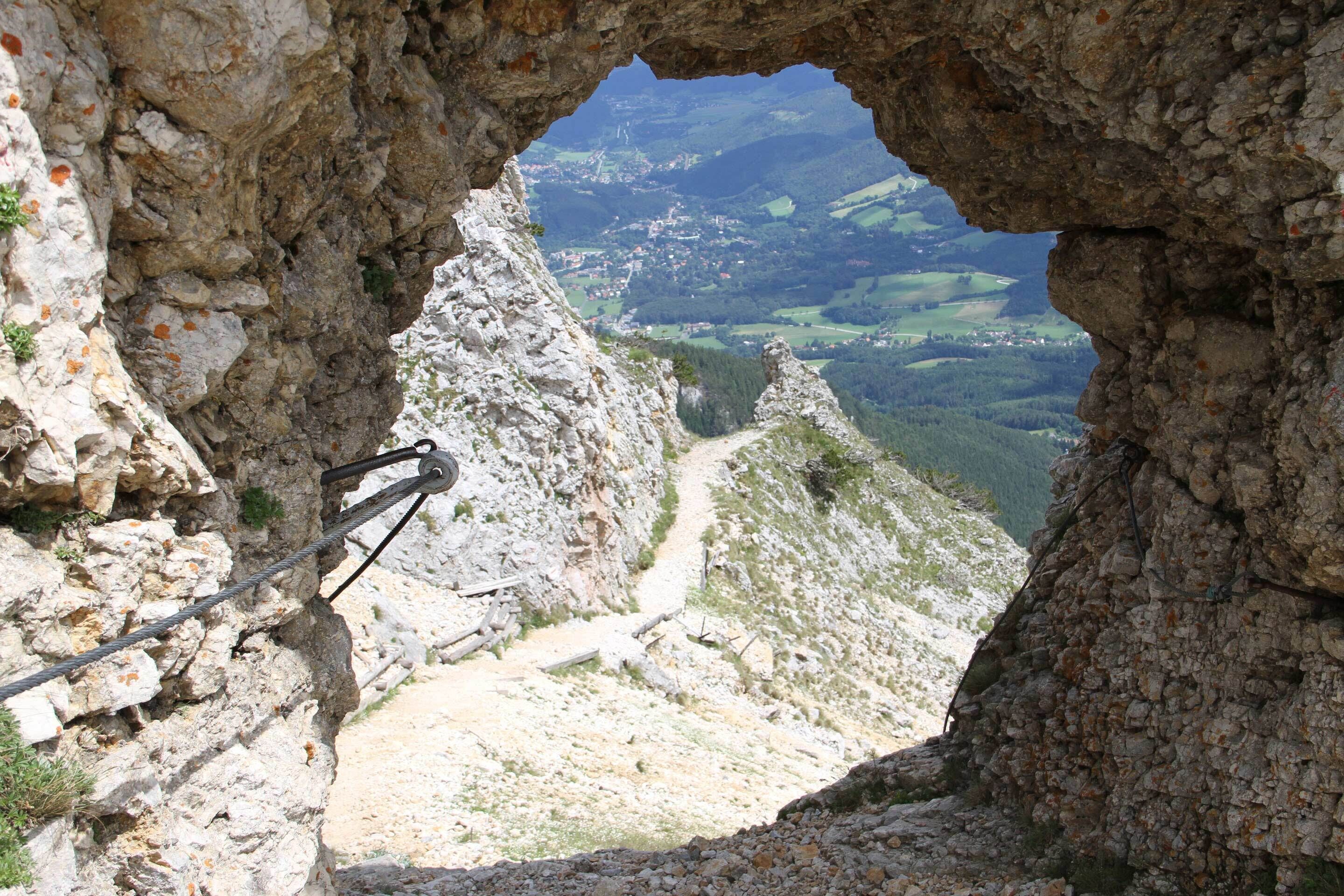A rocky mountain path with a metal railing, a clear view of a valley below, and a distant town.
