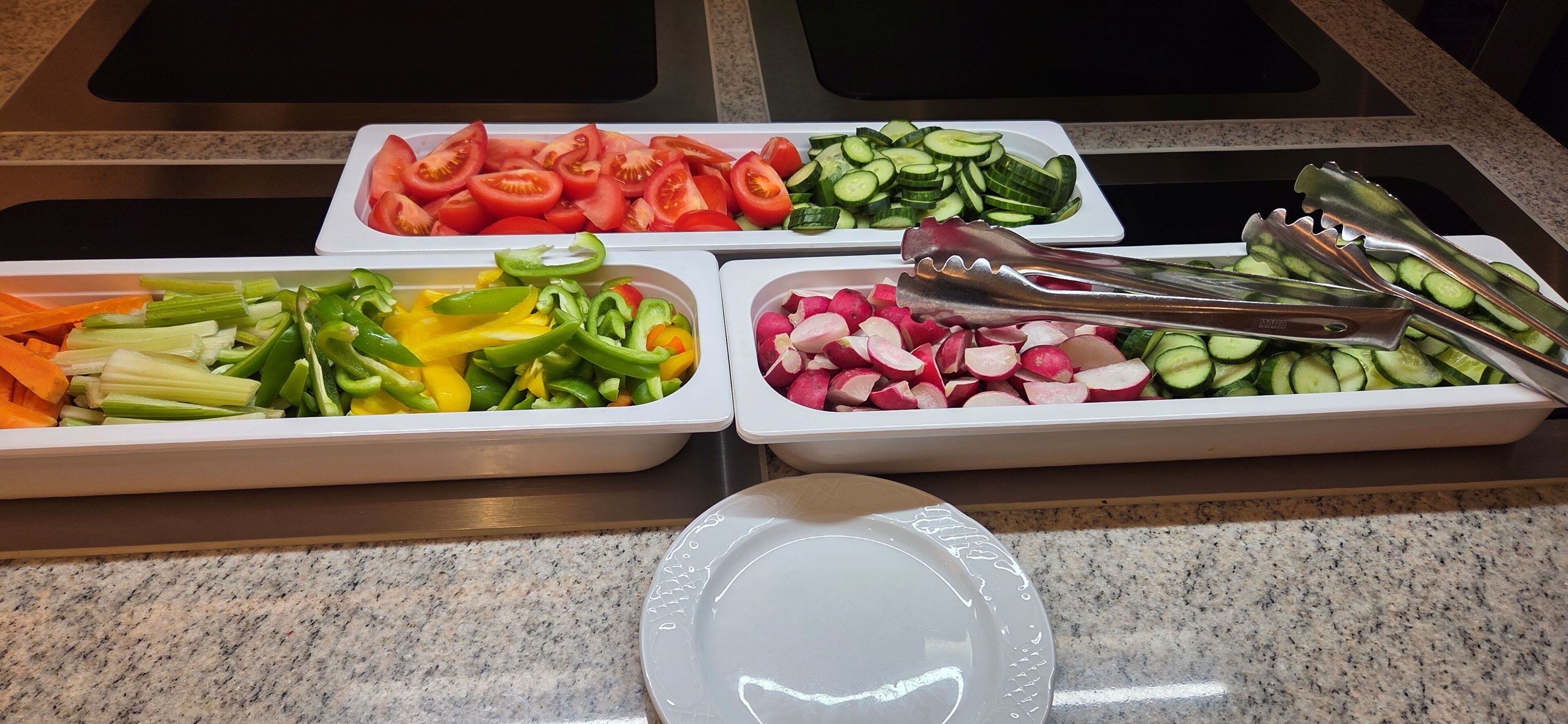 A buffet with various vegetables and a plate with tongs.