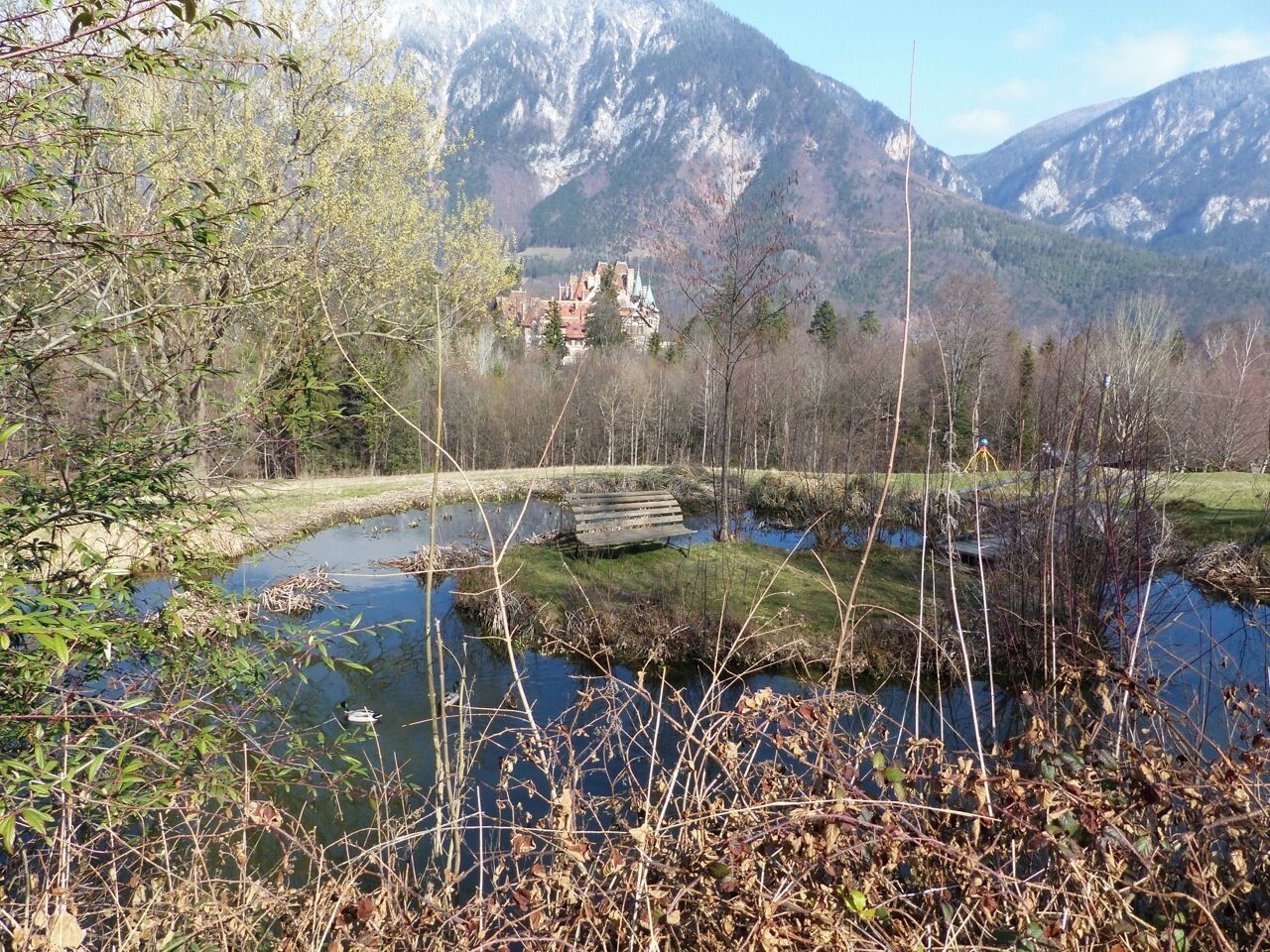 A mountain landscape with a river, a bench, and a building in the background.
