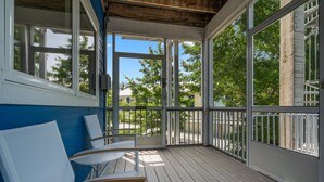 Uma área externa coberta com deck de madeira, cadeiras brancas e grandes janelas com vista para os arredores.
