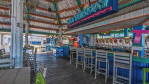 Um bar à beira-mar com um deque de madeira, banquetas e um letreiro de neon que diz "FAT TUESDA".