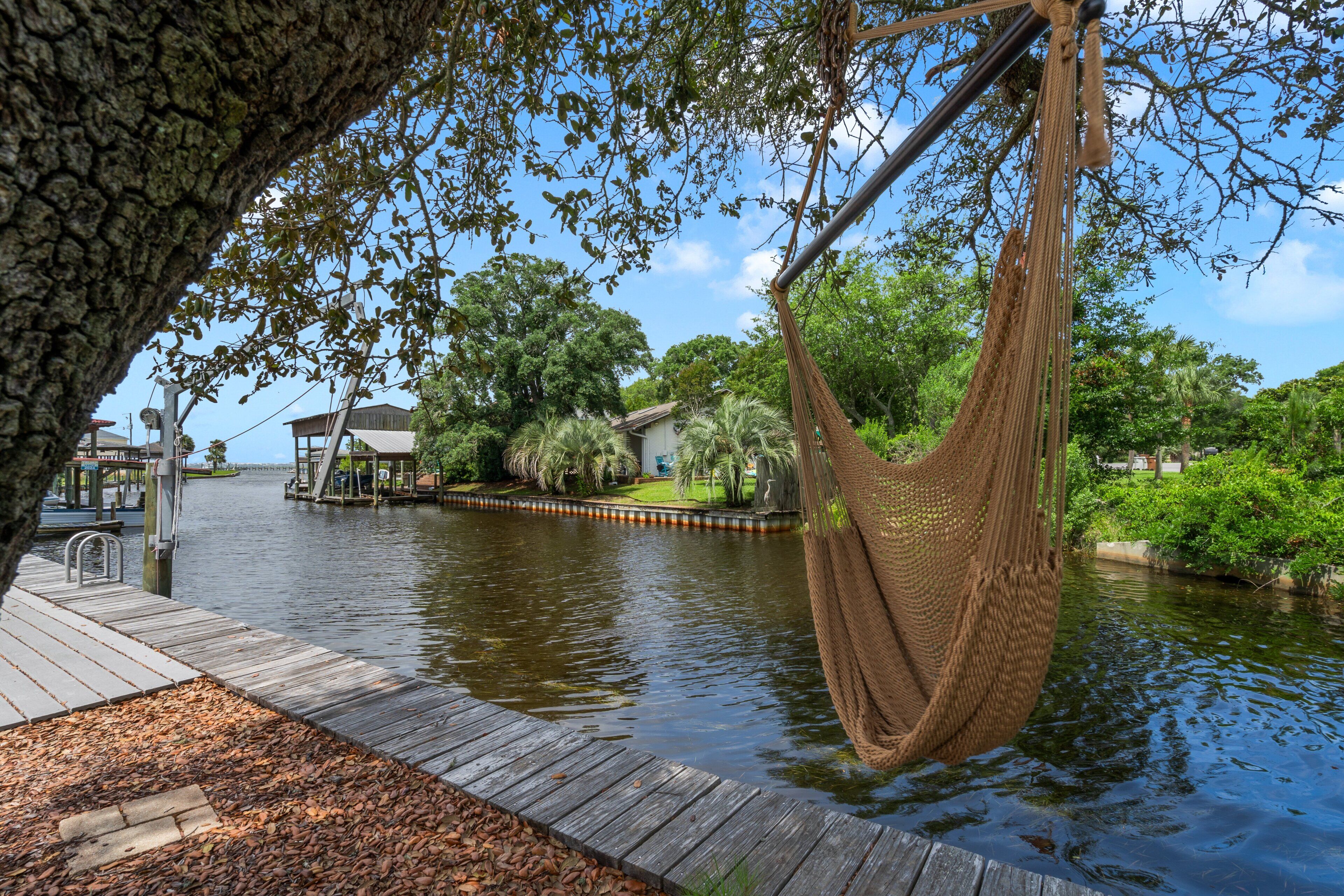 A wooden dock extending over water, a hammock hanging from a tree, and a house with a dock in the background.