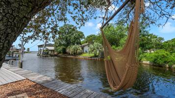A wooden dock extending over water, a hammock hanging from a tree, and a house with a dock in the background.