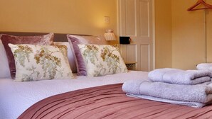 A neatly made bed with floral pillows, a pink bedspread, and a stack of towels. - Templar Mews, heart of town (Cirencester)