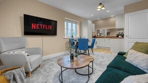 A modern living room with a flat-screen TV, a sofa, a coffee table, and a dining area with chairs and a kitchen in the background. - Discounts for Extended Stays - sleeps 4 (Gloucestershire)