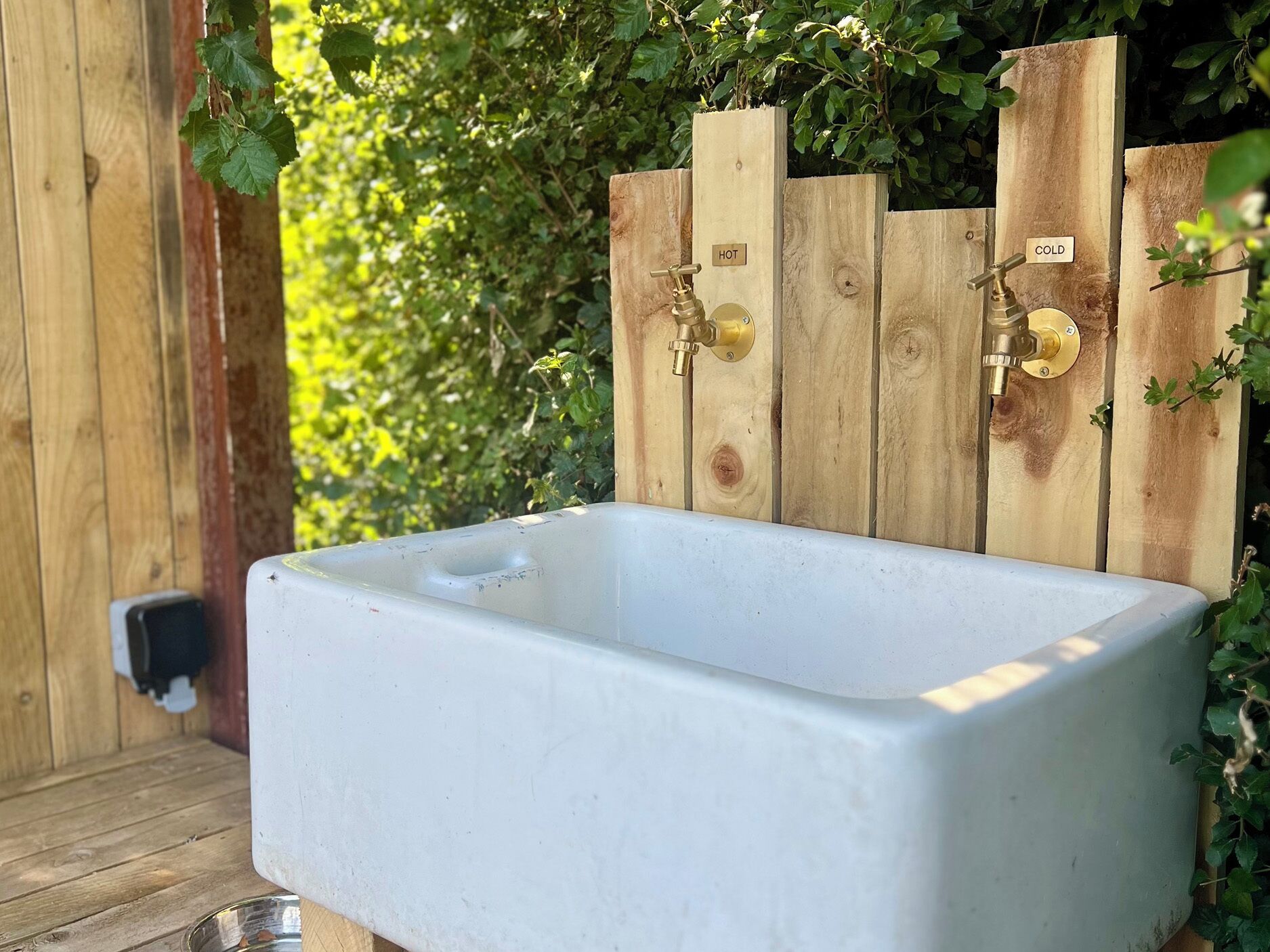 A white rectangular sink with brass faucets, mounted on a wooden fence with 'HOT' and 'COLD' labels.
