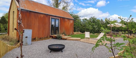 A rustic wooden building with a metal roof, a gravel patio, a black fire pit, and a wooden deck.