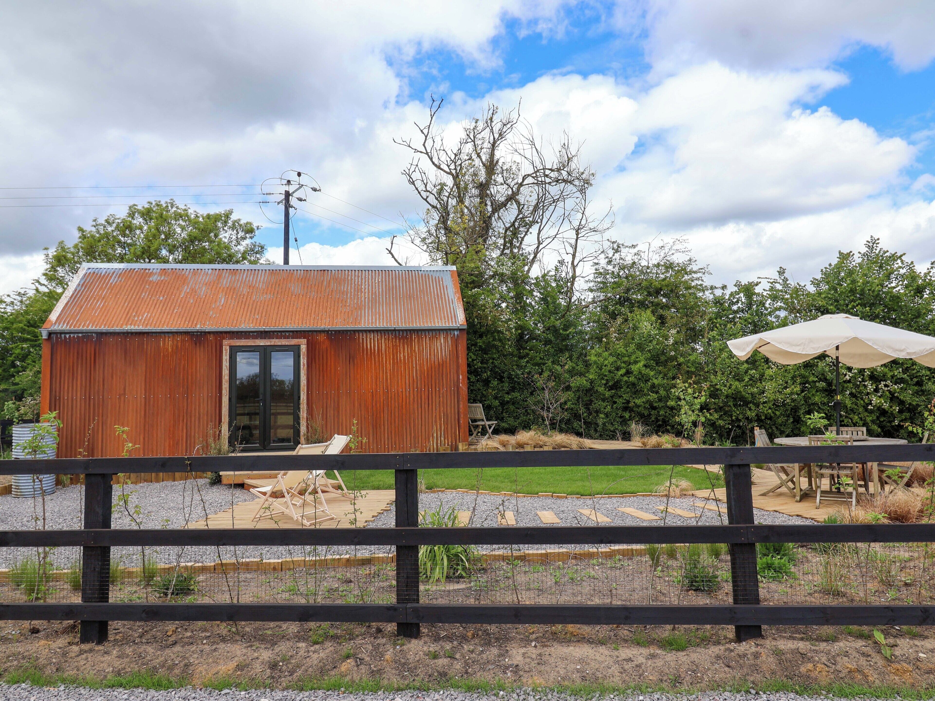 A rustic metal shed with a door, surrounded by a wooden fence, a table with chairs, and an umbrella.