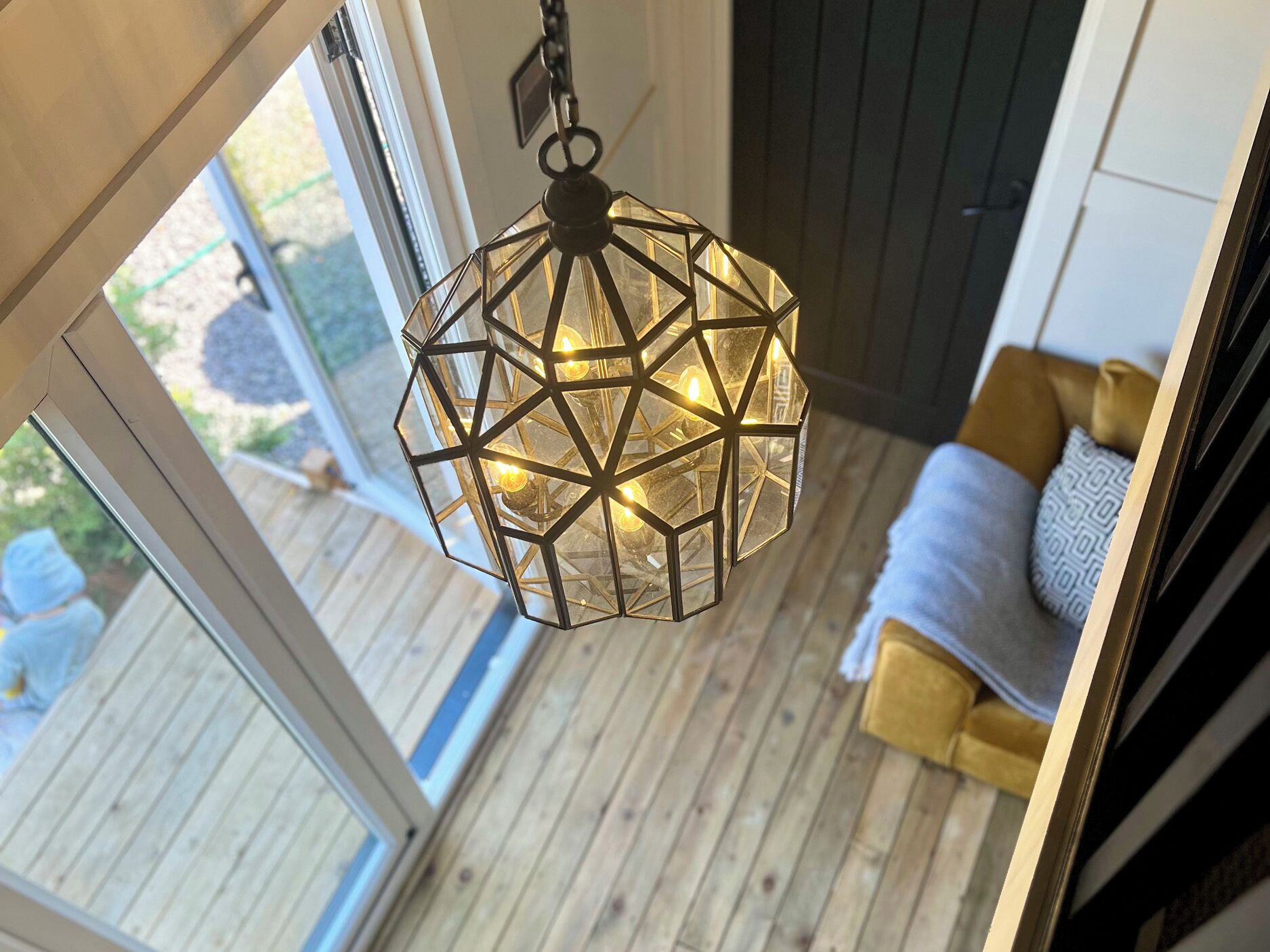 A hexagonal pendant light fixture with multiple bulbs, hanging from a ceiling in a room with wooden flooring and a view of an outdoor deck.