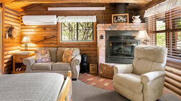 A cozy log cabin interior with a fireplace, a sofa, a chair, and a bed.