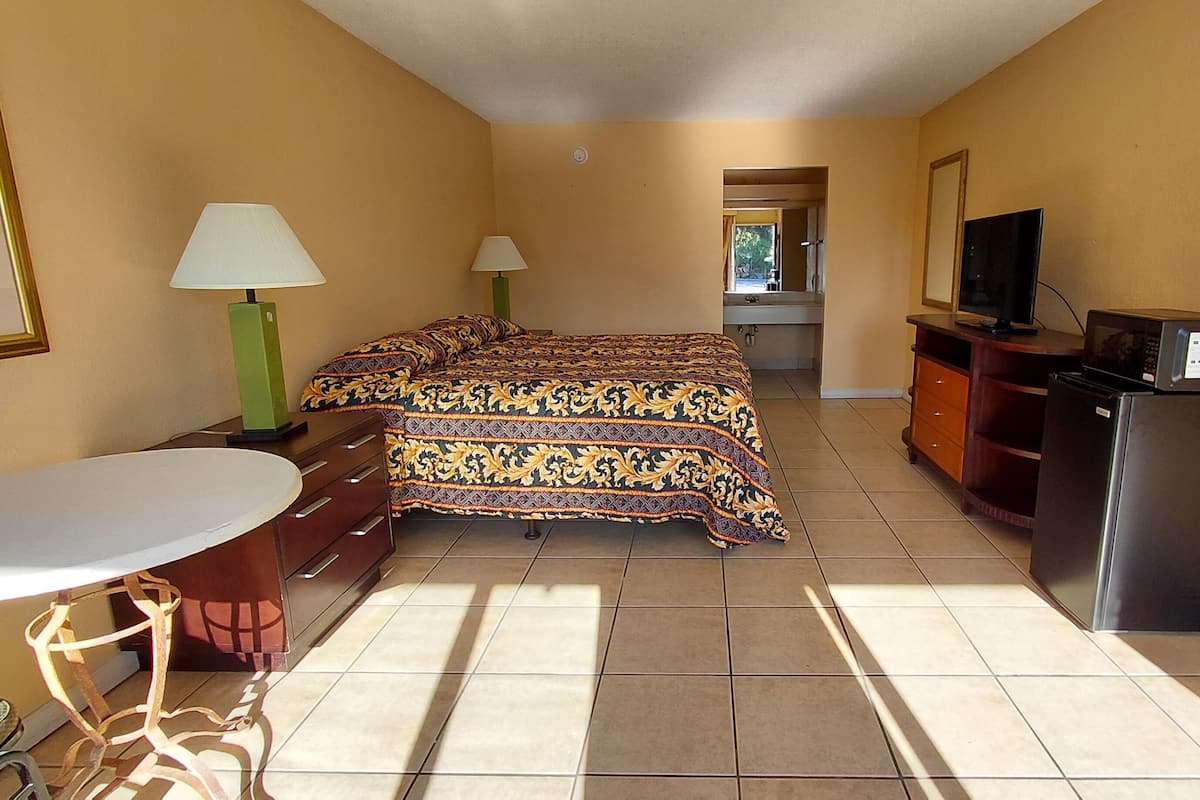 A hotel room with a bed, a desk, a chair, a TV, a refrigerator, and a bathroom visible through an open door.