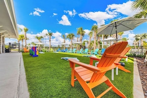 A poolside area with artificial grass, lounge chairs, umbrellas, and palm trees.