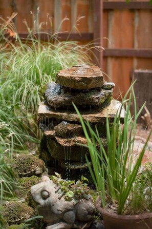 Une fontaine en pierre à plusieurs niveaux, située dans un jardin agrémenté de verdure et entourée d’une clôture en bois.