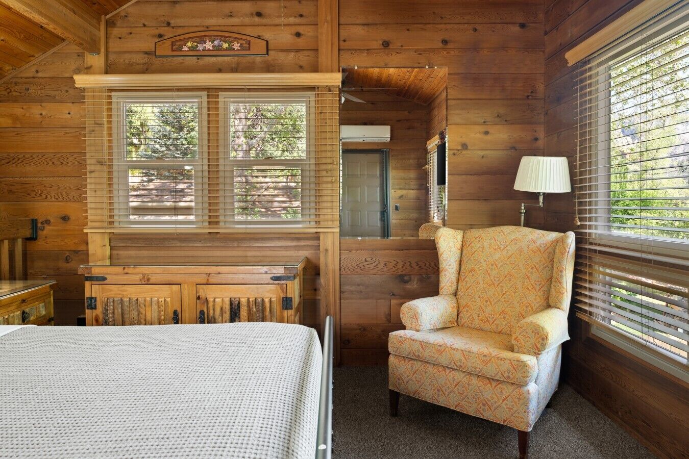 Une pièce de cabane en bois comprenant un lit, une chaise, une lampe et une fenêtre avec des stores.