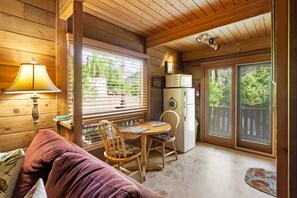 A cozy cabin interior with a wooden ceiling, a dining table with chairs, a refrigerator, and a window with blinds.