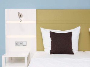 Ein Bett mit weißen und braunen Kissen, ein weißer Nachttisch und eine wandmontierte Leuchte.