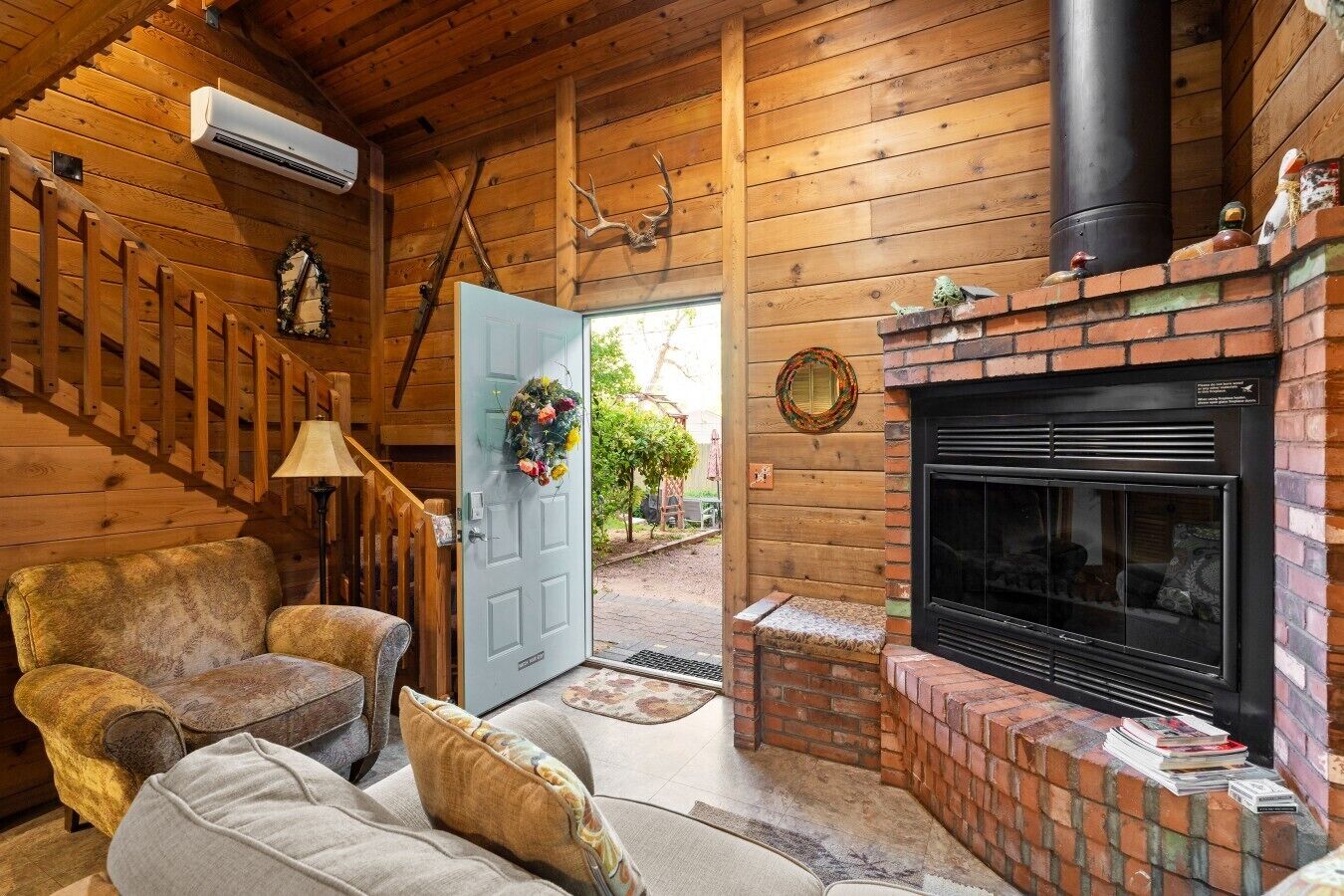 A cozy cabin interior with a fireplace, wooden walls, a staircase, and a door with a floral wreath.