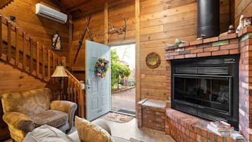 A cozy cabin interior with a fireplace, wooden walls, a staircase, and a door with a floral wreath.